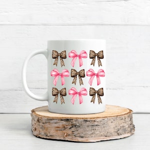 Puede incluir: Una taza de cer&aacute;mica blanca con un patr&oacute;n repetitivo de nueve lazos dispuestos en cuadr&iacute;cula. El dise&ntilde;o alterna entre lazos de color rosa s&oacute;lido y lazos con estampado de leopardo marr&oacute;n, creando una visual equilibrada en la taza.