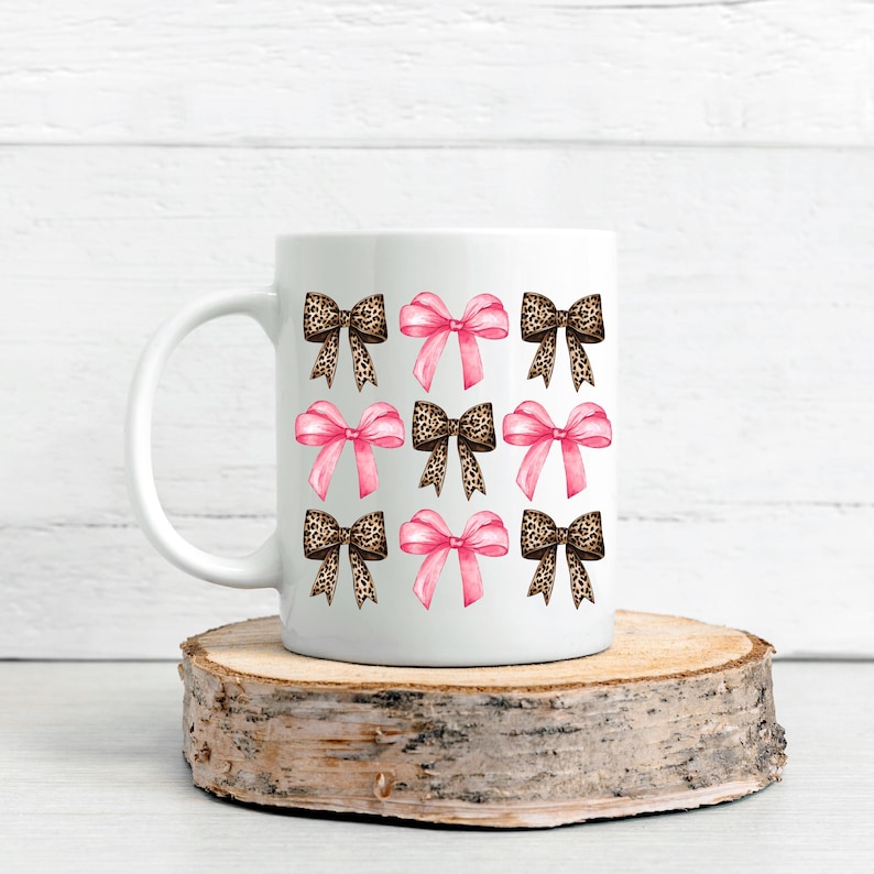 Puede incluir: Una taza de cer&aacute;mica blanca con un patr&oacute;n repetitivo de nueve lazos dispuestos en cuadr&iacute;cula. El dise&ntilde;o alterna entre lazos de color rosa s&oacute;lido y lazos con estampado de leopardo marr&oacute;n, creando una visual equilibrada en la taza.