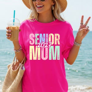 Puede incluir: Una camiseta rosa intenso con las palabras "SENIOR 2027 MOM" en colores pastel. La persona lleva la camiseta, sostiene una bebida y una bolsa de paja, con una playa de fondo.