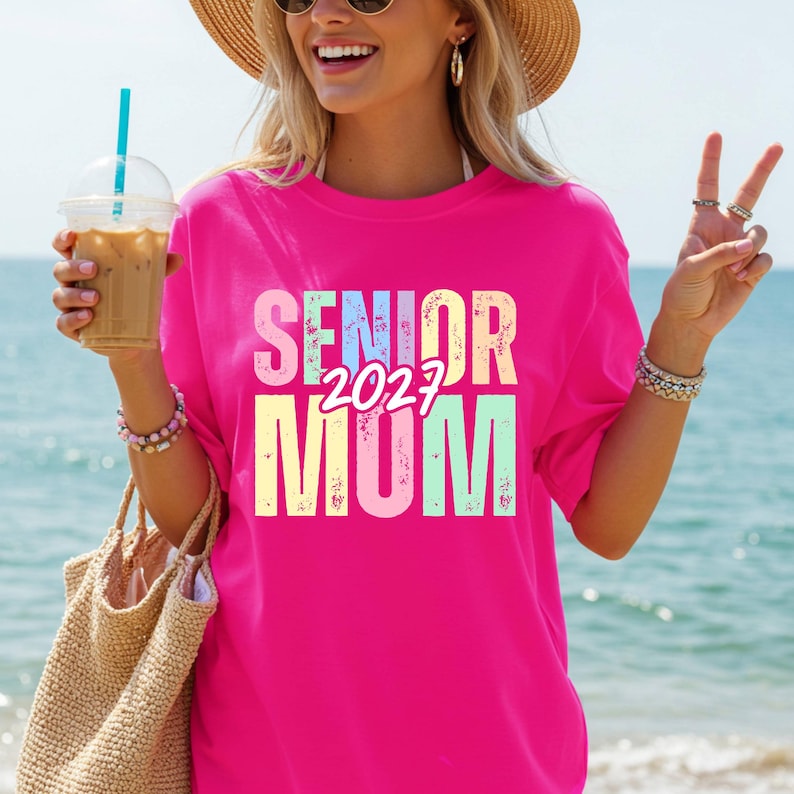 Puede incluir: Una camiseta rosa intenso con las palabras "SENIOR 2027 MOM" en colores pastel. La persona lleva la camiseta, sostiene una bebida y una bolsa de paja, con una playa de fondo.