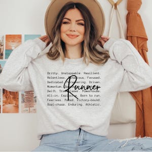 May include: A gray sweatshirt with the text "Gritty. Unstoppable. Resilient. Relentless. Tenacious. Focused. Dedicated. Persevering. Driven. Momentum. Runner. Swift. Trailblazer. Powerhouse. All-in. Explosive. Born to run. Fearless. Paced. Victory-bound. Goal-chase. Enduring. Athletic." printed on the front.
