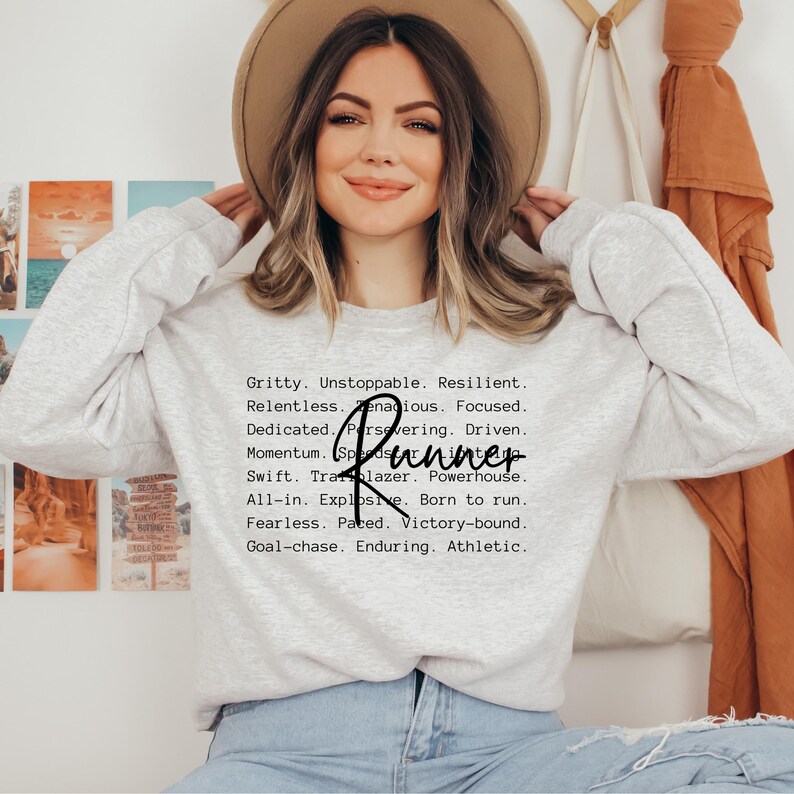 May include: A gray sweatshirt with the text "Gritty. Unstoppable. Resilient. Relentless. Tenacious. Focused. Dedicated. Persevering. Driven. Momentum. Runner. Swift. Trailblazer. Powerhouse. All-in. Explosive. Born to run. Fearless. Paced. Victory-bound. Goal-chase. Enduring. Athletic." printed on the front.