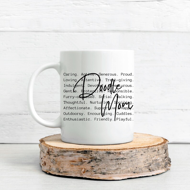 May include: White ceramic coffee mug with a black and white graphic design. The text reads "Doodle Mom" and lists a series of adjectives describing a doodle dog.