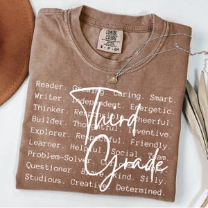 Może przedstawiać: Brązowa koszulka Comfort Colors z napisem "Third Grade" w białym skrypcie. Koszulka wymienia r&oacute;wnież pozytywne cechy, takie jak "Reader", "Writer" i "Thinker". Srebrny naszyjnik z wisiorkiem jest na koszulce.