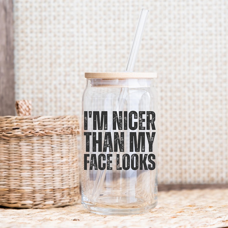 Pu&ograve; includere: Un bicchiere di vetro trasparente con coperchio in legno e cannuccia trasparente. Il bicchiere ha il testo "I'M NICER THAN MY FACE LOOKS" in nero. Il bicchiere &egrave; su una superficie tessuta.