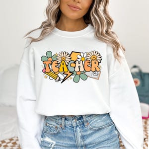 May include: White sweatshirt with a graphic design of the word "TEACHER" in colorful, retro-style letters. The design includes flowers, a lightning bolt, a pencil, and a book with an A+ grade.