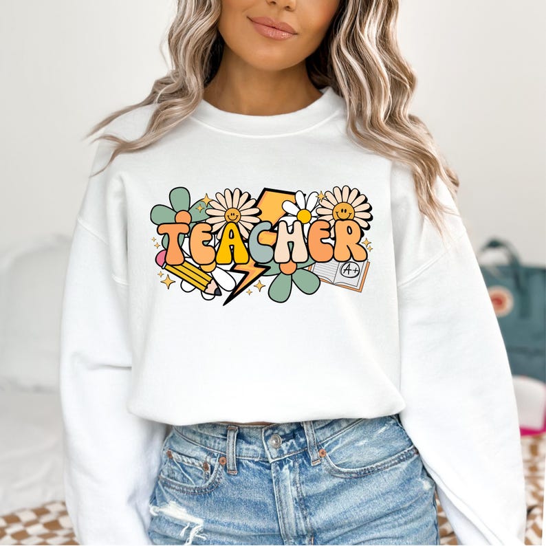 May include: White sweatshirt with a graphic design of the word "TEACHER" in colorful, retro-style letters. The design includes flowers, a lightning bolt, a pencil, and a book with an A+ grade.