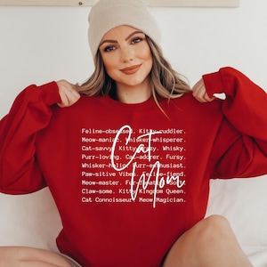 May include: A red sweatshirt with the words "Cat Mom" in white script. The sweatshirt also features a list of cat-related words. The model is wearing a beige beanie.