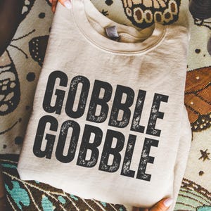 May include: A beige sweatshirt with the words "GOBBLE GOBBLE" printed in large, distressed black letters. The sweatshirt is folded and resting on a patterned surface, suggesting a fall or Thanksgiving theme.