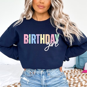 Może przedstawiać: Granatowa bluza z nadrukiem "BIRTHDAY girl" z przodu, w kolorowej, postarzanej czcionce. Słowo "girl" jest napisane kursywą. Bluza ma swobodny styl.