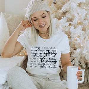 Pu&ograve; includere: Una t-shirt bianca a maniche corte con testo nero che recita: Chocolaty. Flaky. Sticky. Gooey. Chewy. Soft. Cozy. Spiced. Magical. Frosted. Gingery. Sugary. Buttery. Twinkling. Nutty. Sweet. Glazed. Warm. Joy. Crispy. Festive. Crumbly. Christmas Baking.