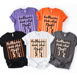 May include: A collection of six t-shirts in various colors: purple, orange, black, white, and gray. Each shirt features the text "Halloween book club" with a bow design and vertical stripes.