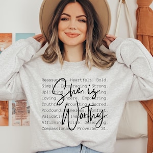 May include: A light gray crewneck sweatshirt featuring black text that reads: Reassuring. Heartfelt. Bold. Simple. Encouraging. Strong. Uplifting. Soulful. Healing. Loving. Sincere. Empowering. Truthful. Grounding. Sacred. Profound. Timeless. Comfort. Validating. Inspiring. Real. Affirming. Pride. Deserving. Compassionate. Proverbs 31. The words She is Worthy are written in a large black script font over the list of words.