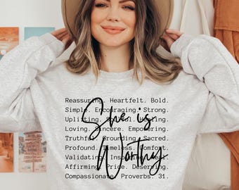 May include: A light grey crewneck sweatshirt featuring black text that reads: Reassuring. Heartfelt. Bold. Simple. Encouraging. Strong. Uplifting. Soulful. Healing. Loving. Sincere. Empowering. Truthful. Grounding. Sacred. Profound. Timeless. Comfort. Validating. Inspiring. Real. Affirming. Pride. Deserving. Compassionate. Proverbs 31. The words She is Worthy are written in a large black script font over the list of words.