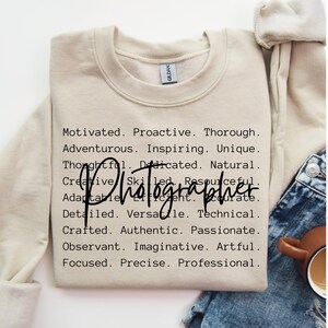 May include: A beige sweatshirt with the text "Motivated. Proactive. Thorough. Adventurous. Inspiring. Unique. Thoughtful. Dedicated. Natural. Creative. Skilled. Resourceful. Adaptable. Efficient. Accurate. Detailed. Versatile. Technical. Crafted. Authentic. Passionate. Observant. Imaginative. Artful. Focused. Precise. Professional." and the word "Photographer" in large, bold letters.