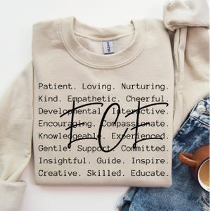 May include: A beige sweatshirt with the words "Patient. Loving. Nurturing. Kind. Empathetic. Cheerful. Developmental. Interactive. Encouraging. Compassionate. Knowledgeable. Experienced. Gentle. Support. Committed. Insightful. Guide. Inspire. Creative. Skilled. Educate." printed on the front. The word "HOT" is printed in large letters in the centre of the sweatshirt.