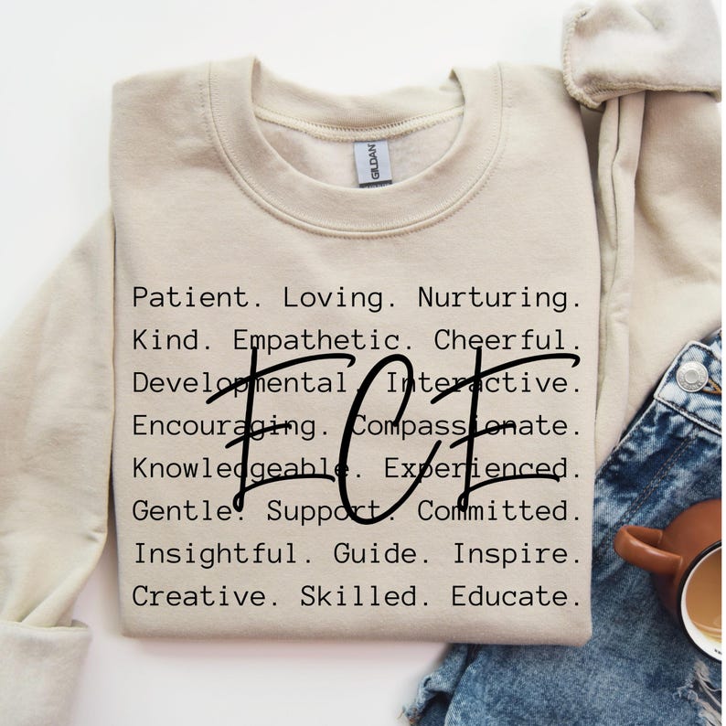 May include: A beige sweatshirt with the words "Patient. Loving. Nurturing. Kind. Empathetic. Cheerful. Developmental. Interactive. Encouraging. Compassionate. Knowledgeable. Experienced. Gentle. Support. Committed. Insightful. Guide. Inspire. Creative. Skilled. Educate." printed on the front. The word "HOT" is printed in large letters in the centre of the sweatshirt.