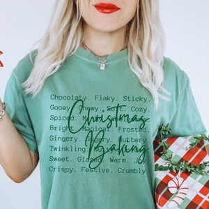 Pu&ograve; includere: Una t-shirt a maniche corte color verde salvia con testo stampato in nero che elenca vari aggettivi come Chocolaty, Flaky, Sticky e altri, con la scritta in corsivo verde scuro Christmas Baking sovrapposta al centro della t-shirt.