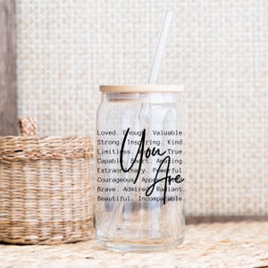 May include: A clear glass jar with a wooden lid and a straw. The jar has a black inscription that reads "Loved. Enough. Valuable. Strong. Inspiring. Kind. Limitless. True. Capable. Smart. Amazing. Extraordinary. Powerful. Courageous. Appreciated. Brave. Admired. Radiant. Beautiful. Incomparable. You Are."
