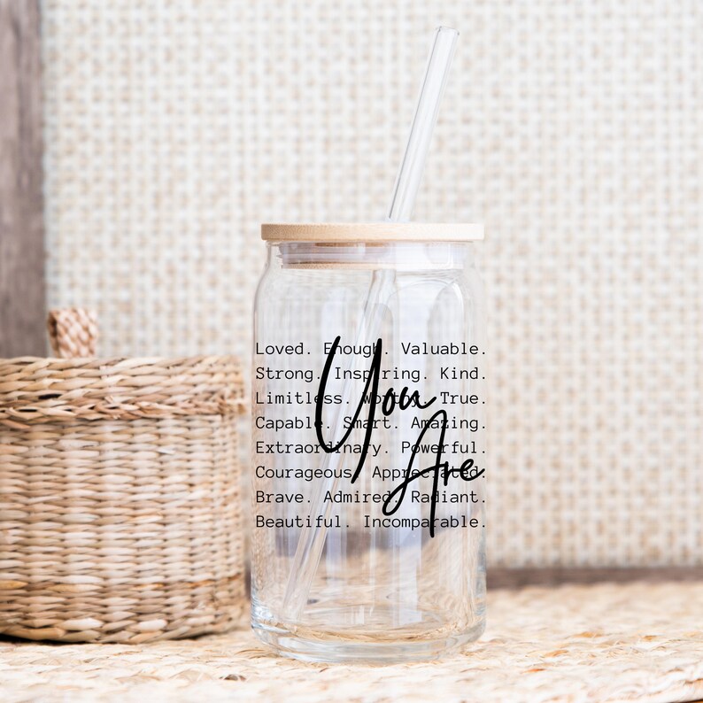 May include: A clear glass jar with a wooden lid and a straw. The jar has a black inscription that reads "Loved. Enough. Valuable. Strong. Inspiring. Kind. Limitless. True. Capable. Smart. Amazing. Extraordinary. Powerful. Courageous. Appreciated. Brave. Admired. Radiant. Beautiful. Incomparable. You Are."