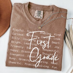 May include: A brown Comfort Colors t-shirt with the words "First Grade" in white script. The shirt is adorned with a list of positive adjectives. A silver necklace with a pendant is draped across the shirt.