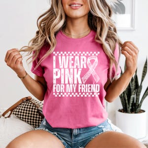 May include: A pink t-shirt with the text "I WEAR PINK FOR MY FRIEND" in white, along with a white ribbon graphic. The shirt has a checkered border at the top and bottom of the text.