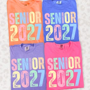May include: Four crew neck t-shirts in orange, blue, purple, and pink. Each shirt features the text SENIOR 2027 printed in a multi-colored, distressed font with letters and numbers in shades of pink, blue, yellow, and green across the chest.