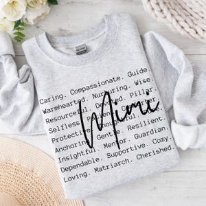 May include: Grey sweatshirt with the word "Mimi" written in black cursive. The text below the name lists positive attributes such as caring, compassionate, guide, warmhearted, nurturing, wise, resourceful, devoted, pillar, selfless, patient, thoughtful, gentle, resilient, anchoring, mentor, guardian, insightful, dependable, supportive, cosy, loving, and matriarch.