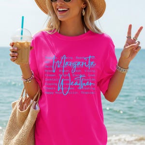 May include: A bright pink t-shirt with the words "Margarita Weather" in blue script. The shirt features a list of related words. The person in the image is wearing sunglasses and a straw hat, holding a drink and a woven bag.