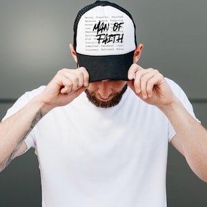 Op de afbeelding: Een zwart-witte truckerpet met de tekst "MAN OF FAITH" in het midden. De pet heeft ook een lijst met woorden zoals "Devout", "Prayerful" en "Anointed".
