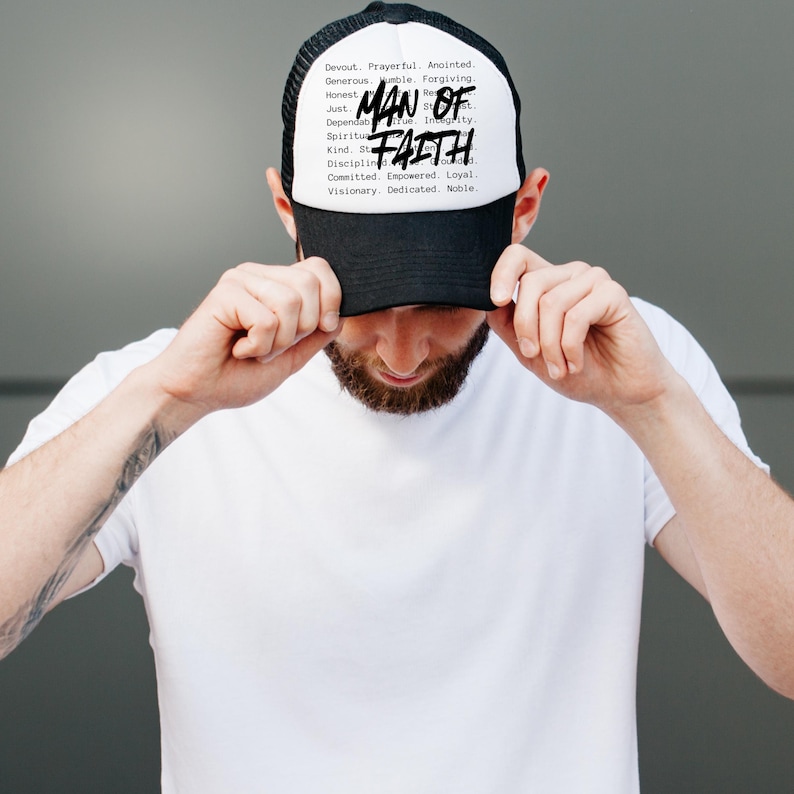 Op de afbeelding: Een zwart-witte truckerpet met de tekst "MAN OF FAITH" in het midden. De pet heeft ook een lijst met woorden zoals "Devout", "Prayerful" en "Anointed".
