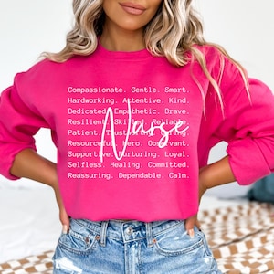 K&ouml;nnte beinhalten: Ein rosa Sweatshirt mit dem Wort "Krankenschwester" in wei&szlig;er Schrift. Das Sweatshirt hat eine Liste positiver Attribute, die mit dem Wort "Krankenschwester" verbunden sind, darunter mitf&uuml;hlend, sanft, klug, flei&szlig;ig, aufmerksam, freundlich, engagiert, empathisch, mutig, belastbar, geschickt, zuverl&auml;ssig, geduldig, vertrauensw&uuml;rdig, f&uuml;rsorglich, einfallsreich, Held, aufmerksam, unterst&uuml;tzend, pflegend, loyal, selbstlos, heilend, engagiert, beruhigend, zuverl&auml;ssig und ruhig.