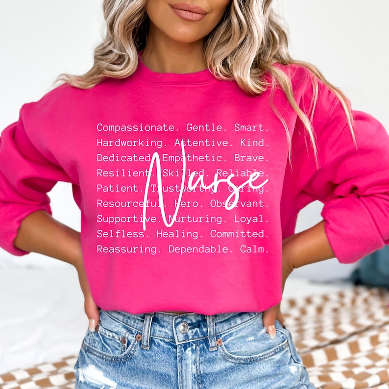 K&ouml;nnte beinhalten: Ein rosa Sweatshirt mit dem Wort "Krankenschwester" in wei&szlig;er Schrift. Das Sweatshirt hat eine Liste positiver Attribute, die mit dem Wort "Krankenschwester" verbunden sind, darunter mitf&uuml;hlend, sanft, klug, flei&szlig;ig, aufmerksam, freundlich, engagiert, empathisch, mutig, belastbar, geschickt, zuverl&auml;ssig, geduldig, vertrauensw&uuml;rdig, f&uuml;rsorglich, einfallsreich, Held, aufmerksam, unterst&uuml;tzend, pflegend, loyal, selbstlos, heilend, engagiert, beruhigend, zuverl&auml;ssig und ruhig.