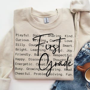 May include: A beige sweatshirt with the words "First Grade" in a handwritten font. The sweatshirt is printed with a list of positive adjectives such as "Playful," "Kind," and "Smart."