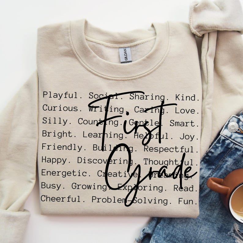 May include: A beige sweatshirt with the words "First Grade" in a handwritten font. The sweatshirt is printed with a list of positive adjectives such as "Playful," "Kind," and "Smart."