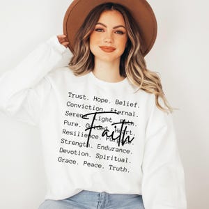 Puede incluir: Sudadera blanca con la palabra "Fe" en letras negras y una lista de palabras asociadas a la fe, incluyendo confianza, esperanza, creencia, convicci&oacute;n, eterno, sereno, luz, camino, puro, guiado, coraz&oacute;n, resiliencia, prop&oacute;sito, fuerza, resistencia, devoci&oacute;n, espiritual, gracia, paz y verdad.