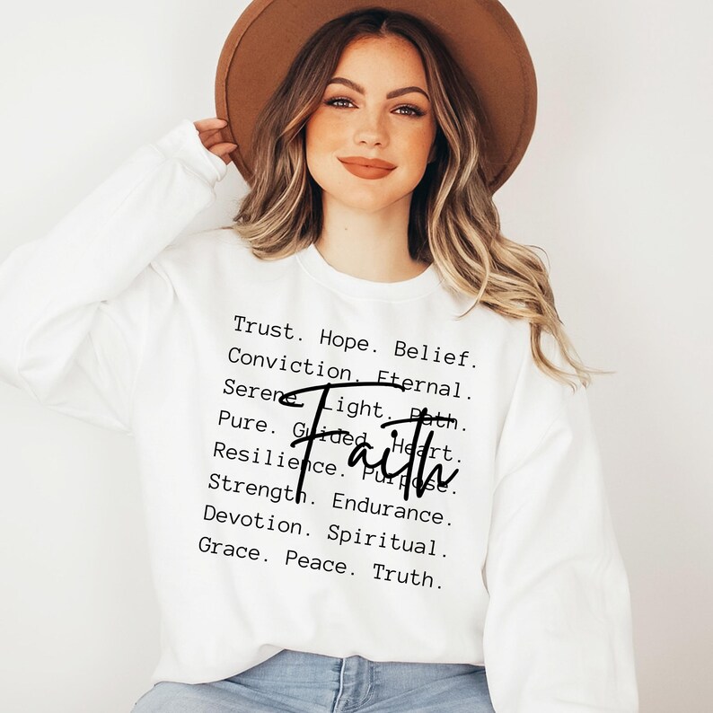 Puede incluir: Sudadera blanca con la palabra "Fe" en letras negras y una lista de palabras asociadas a la fe, incluyendo confianza, esperanza, creencia, convicci&oacute;n, eterno, sereno, luz, camino, puro, guiado, coraz&oacute;n, resiliencia, prop&oacute;sito, fuerza, resistencia, devoci&oacute;n, espiritual, gracia, paz y verdad.