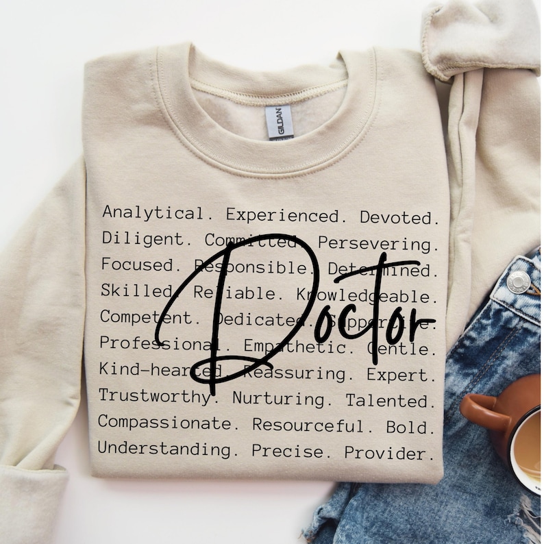 May include: A beige sweatshirt with the word "Doctor" in large black letters surrounded by a list of positive attributes, such as "analytical", "experienced", "devoted", "diligent", "committed", "persevering", "focused", "responsible", "determined", "skilled", "reliable", "knowledgeable", "competent", "dedicated", "professional", "empathetic", "gentle", "kind-hearted", "reassuring", "expert", "trustworthy", "nurturing", "talented", "compassionate", "resourceful", "bold", "understanding", "precise", and "provider".