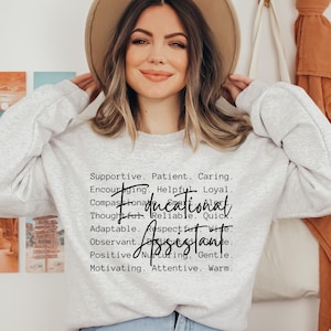 Op de afbeelding: Een lichtgrijs sweatshirt met ronde hals en de zwarte tekst Educational Assistant in een cursief lettertype, omringd door een lijst met beschrijvende woorden zoals Supportive, Patient, Caring, Encouraging, Helpful, Loyal, Compassionate, Smart, Alert, Thoughtful, Reliable, Quick, Adaptable, Respectful, Wise, Observant, Dedicated, Positive, Nurturing, Gentle, Motivating, Attentive en Warm.