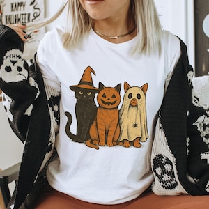K&ouml;nnte beinhalten: Wei&szlig;es T-Shirt mit einem Halloween-Motiv von drei Cartoon-Katzen. Eine ist schwarz mit einem Hexenhut, eine andere ist orange mit einem K&uuml;rbisgesicht und die dritte ist eine Geisterkatze. Die Worte "HAPPY HALLOWEEN" sind im Hintergrund zu sehen.