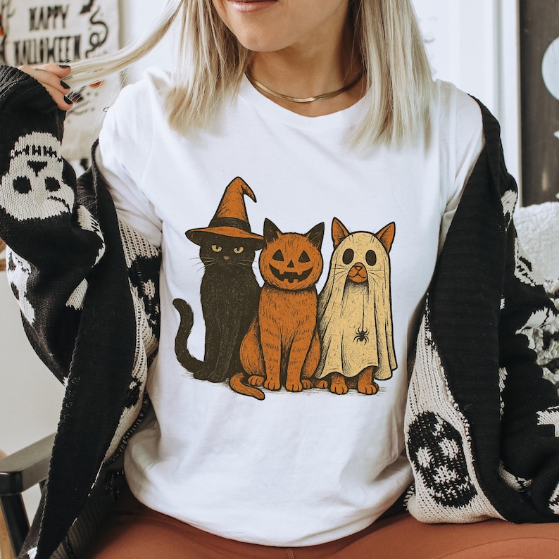 K&ouml;nnte beinhalten: Wei&szlig;es T-Shirt mit einem Halloween-Motiv von drei Cartoon-Katzen. Eine ist schwarz mit einem Hexenhut, eine andere ist orange mit einem K&uuml;rbisgesicht und die dritte ist eine Geisterkatze. Die Worte "HAPPY HALLOWEEN" sind im Hintergrund zu sehen.