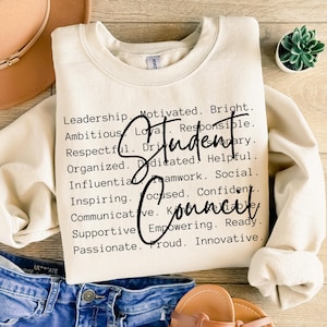 May include: A cream-colored crewneck sweatshirt featuring black text that lists various positive traits like Leadership, Motivated, Bright, Ambitious, Loyal, Responsible, Respectful, Driven, Organized, Dedicated, Helpful, Influential, Teamwork, Social, Inspiring, Focused, Confident, Communicative, Knowledgeable, Supportive, Empowering, Ready, Passionate, Proud, and Innovative, with the words Student Council written in a large black script font across the center.