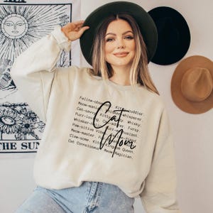 May include: Beige sweatshirt with the words "Cat Mom" in a script font. The sweatshirt has a list of cat-related words in a vertical column. The person in the image is wearing a dark green hat and blue jeans.