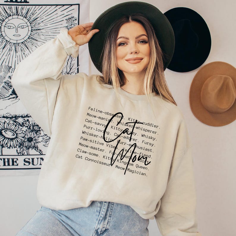 May include: Beige sweatshirt with the words "Cat Mom" in a script font. The sweatshirt has a list of cat-related words in a vertical column. The person in the image is wearing a dark green hat and blue jeans.