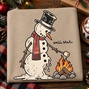 May include: A tan sweatshirt featuring a graphic of a melting snowman wearing a black top hat and a red scarf. The snowman holds a marshmallow over a campfire with the text well, shit... printed in black next to the snowman's orange carrot nose.