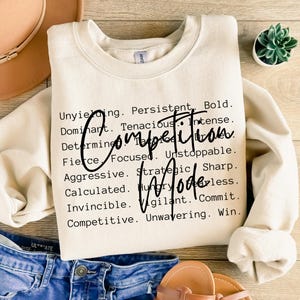 May include: A cream-colored crewneck sweatshirt featuring black text that lists various adjectives including Unyielding, Persistent, Bold, Dominant, Tenacious, Intense, Determined, Fierce, Focused, Unstoppable, Aggressive, Strategic, Sharp, Calculated, Hungry, Fearless, Invincible, Vigilant, Commit, Competitive, Unwavering, Win, with the words Competition Mode written in a large black script font across the center.