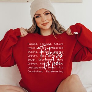 May include: A red sweatshirt with the words "Fitness Mode" and motivational words in white. The sweatshirt is being held up by a person wearing a beige beanie.