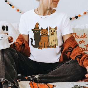 K&ouml;nnte beinhalten: Wei&szlig;es T-Shirt mit einem Halloween-Design, das drei Katzen zeigt. Eine Katze tr&auml;gt einen Hexenhut, eine andere ein geschnitztes K&uuml;rbisgesicht und die dritte ist ein Geist. Das Shirt ist Teil einer Halloween-Kollektion.