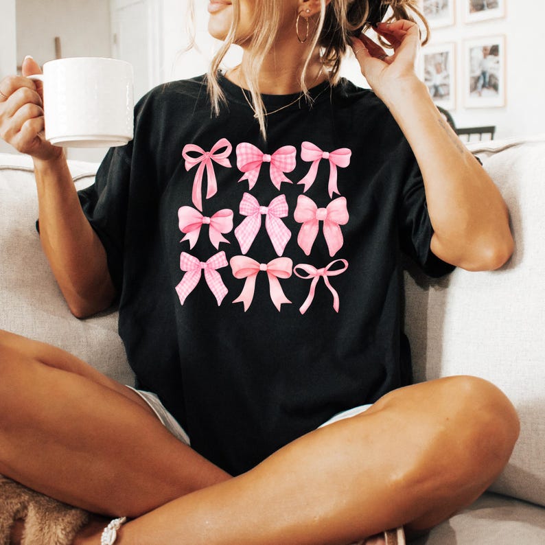 Puede incluir: Camiseta negra con un dise&ntilde;o de nueve lazos rosas en una cuadr&iacute;cula. Los lazos presentan diferentes patrones, lisos y de cuadros. La camiseta tiene un corte holgado y de gran tama&ntilde;o.