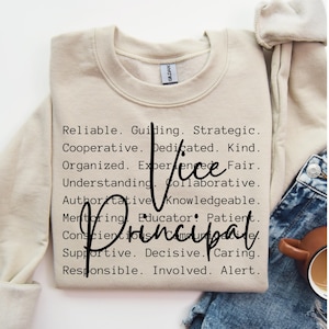Op de afbeelding: Een beige sweatshirt met de tekst "Vice Principal" in zwarte letters. De tekst wordt omringd door een lijst met positieve eigenschappen, zoals "betrouwbaar", "leidend", "strategisch", "samenwerkend", "toegewijd", "vriendelijk", "georganiseerd", "ervaren", "eerlijk", "begripvol", "samenwerkend", "gezaghebbend", "kennisrijk", "mentoring", "opvoeder", "geduldig", "zorgvuldig", "ondersteunend", "beslissend", "zorgzaam", "verantwoordelijk", "betrokken" en "alert".